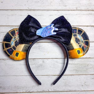 May include: A black headband with attached mouse ears. The ears feature a gold and black design with a window graphic. A black bow with a light blue spaceship charm is centered on the headband. The ears are approximately 4 inches in diameter.
