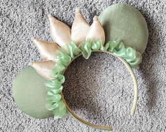Può includere: Una fascia decorativa con accenti verdi e beige. La fascia presenta due grandi orecchie rotonde verde salvia e una serie di abbellimenti beige a forma di foglia con accenti glitterati. Un tessuto verde chiaro arruffato adorna la base delle foglie.