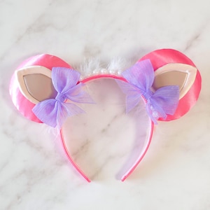 Puede incluir: Diadema de orejas de ratón de satén rosa con lazos lavanda y detalles de piel sintética. Las orejas tienen un detalle interior de color beige claro. La diadema está adornada con plumas blancas y pequeñas perlas.