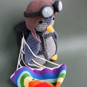 Pode incluir: Boneca de pinguim de crochê usando um chapéu de aviador marrom e óculos, com uma mochila cinza e um paraquedas arco-íris. O pinguim tem um bico amarelo e é feito de fio cinza e azul.