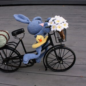 Puede incluir: Un conejito de crochet azul montando una bicicleta negra con una cesta llena de flores de crochet blancas. El conejito sostiene un pato de crochet amarillo.