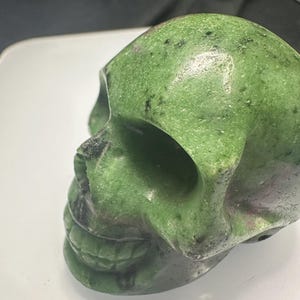 Könnte beinhalten: Ein grüner und schwarzer geschnitzter Schädel aus Stein. Der Schädel hat ein detailliertes Gesicht mit einem breiten Mund und sichtbaren Zähnen.