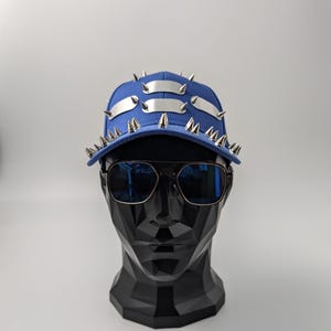 Puede incluir: Una gorra de béisbol azul con pinchos de metal plateado y dos bandas de metal plateado en la parte delantera. La gorra está en una cabeza de maniquí negra.