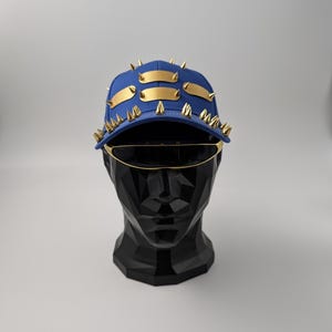 Puede incluir: Una gorra de béisbol azul con pinchos dorados y detalles de metal dorado. La gorra está en una cabeza de maniquí negra.