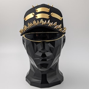 Op de afbeelding: Een zwarte baseballpet met gouden spikes en drie gouden banden over de voorkant. De pet zit op een zwart mannequinhoofd.
