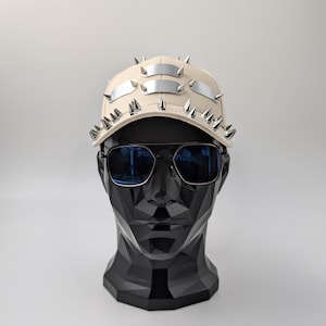 Könnte beinhalten: Eine beige Baseballkappe mit silbernen Metallakzenten und Spikes. Die Kappe sitzt auf einem schwarzen Mannequin-Kopf, der eine blaue Sonnenbrille trägt.
