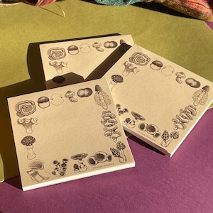 Może przedstawiać: Trzy brązowe notesy z ozdobnym motywem grzybów. Notesy są puste i gotowe do użycia do pisania lub rysowania. Notesy leżą na fioletowym tle.