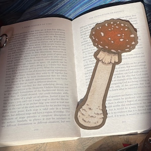 Könnte beinhalten: Ein handgefertigter Lesezeichen in Pilzform mit braun-weiß geflecktem Hut und beigem Stiel. Das Lesezeichen liegt auf einem offenen Buch mit Text und einer Schere.
