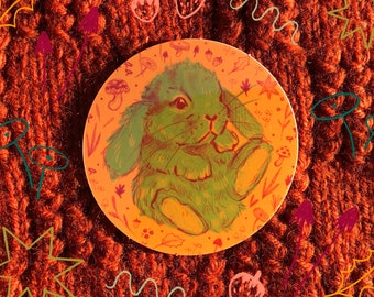 May include: A round sticker with a green and yellow cartoon bunny rabbit on a peach background. The bunny is sitting with its paws tucked in and has a cute expression. The background is filled with small floral and mushroom illustrations.
