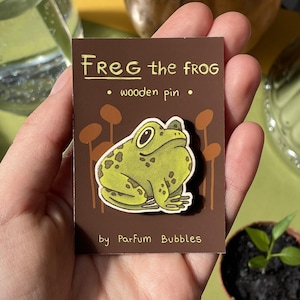 May include: A wooden pin featuring a cartoon illustration of a green frog with brown spots. The pin is titled "Freg the frog" and says "wooden pin" below the image. The pin is made by "Parfum Bubbles".