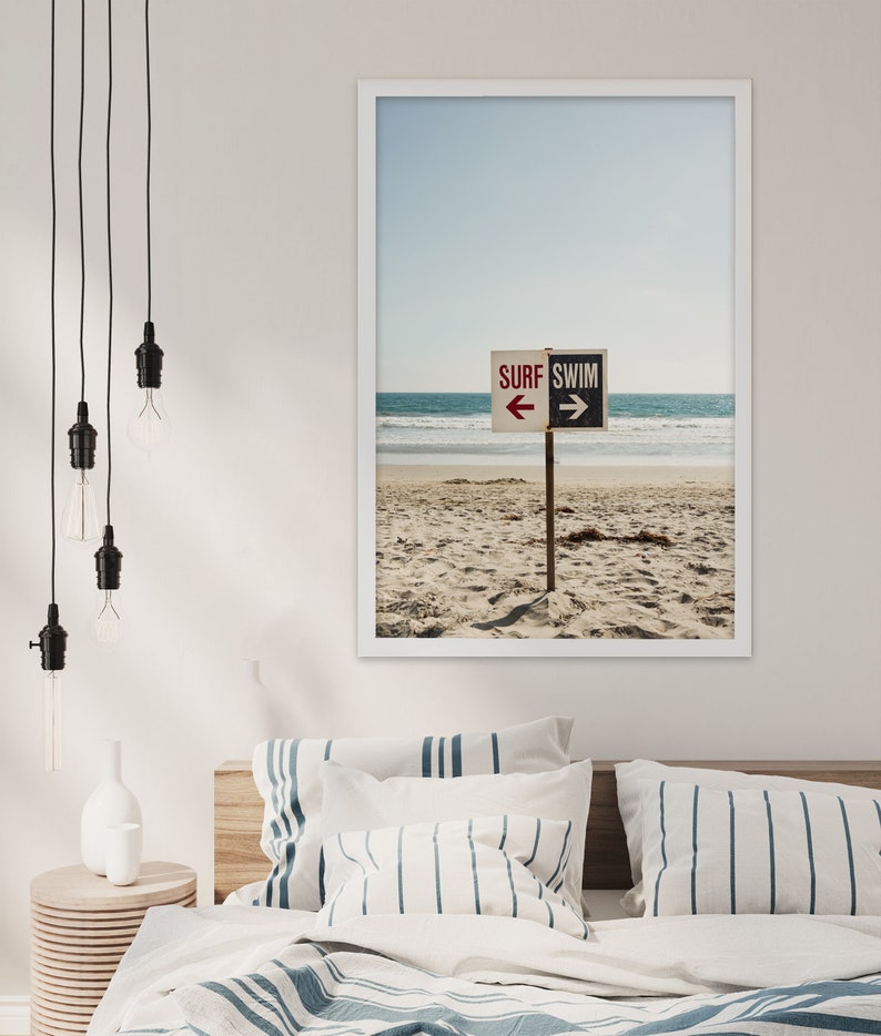 Op de afbeelding: Een ingelijste foto van een strand met een bord waarop "SURF SWIM" staat met pijlen die in tegengestelde richtingen wijzen. Het bord staat op een houten paal en is in het zand geplaatst. Het strand is leeg en de lucht is blauw.