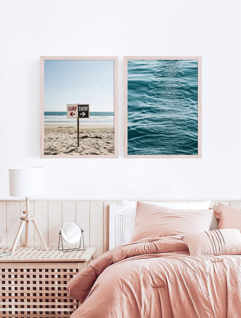 Op de afbeelding: Twee ingelijste prints van strandscenes. De eerste print toont een strand met een bord waarop "Surf Swim" staat, met pijlen die in tegengestelde richtingen wijzen. De tweede print toont een close-up van blauwe oceaan golven.