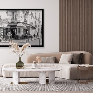 May include: A black and white photograph of a Parisian street scene with a cafe called "Le Hibou" and a sign that says "Patisserie". The photo is framed in black and hanging on a white wall above a beige sofa with a marble coffee table and two gold side tables.
