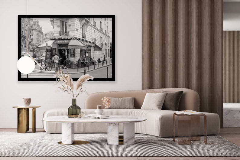 May include: A black and white photograph of a Parisian street scene with a cafe called "Le Hibou" and a sign that says "Patisserie". The photo is framed in black and hanging on a white wall above a beige sofa with a marble coffee table and two gold side tables.