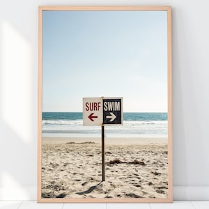 Op de afbeelding: Een strandscene met een bord waarop "SURF" staat met een pijl naar links en "SWIM" met een pijl naar rechts. Het bord staat op een houten paal en is in het zand geplaatst. De zee is op de achtergrond.