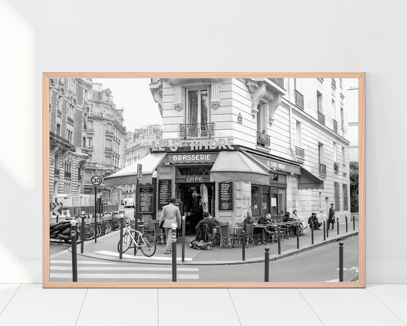 May include: Black and white photograph of a Parisian cafe called "Le Saint-Andre" with a sign that says "Brasserie" and "Cafe".  People are sitting outside the cafe at tables and chairs.