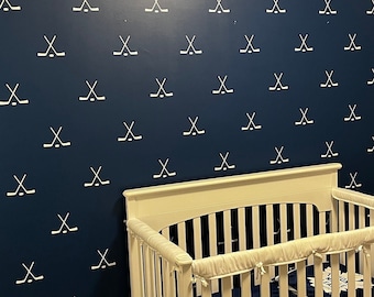 May include: A dark blue wall featuring a repeating white pattern of crossed hockey sticks. In front of the wall sits a white wooden crib with a blue and white patterned blanket inside, and a white bookshelf filled with books stands to the right.