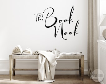 Puede incluir: Un banco de madera blanco con una manta blanca drapeada sobre él. Un libro abierto está en el banco. La pared detrás del banco tiene una calcomanía de vinilo negro que dice "The Book Nook".