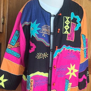 May include: A long-sleeved jacket featuring a vibrant, multi-colored geometric and star-patterned print on a black base. The garment includes bright pink, orange, blue, and yellow accents, a round neckline, and orange toggle-style button closures.