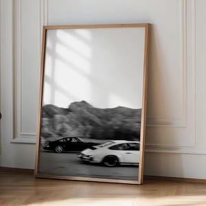 以下が含まれることがあります： 木製のフレームに入った、2台のレーシングカーがレースをしている白黒写真。車は動きを表現するためにぼやけており、背景には山脈が写っています。フレームは明るい木の色です。