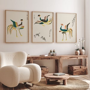 May include: Three framed prints of stylized bird illustrations in shades of green, blue, yellow, and brown. The prints are hung on a white wall above a wooden table and a white armchair.