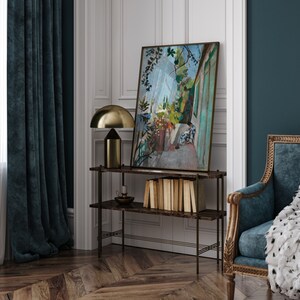 May include: A black metal shelf unit with a painting of a garden scene hanging above it. The shelf unit has three shelves and is standing on a wood floor. A gold table lamp is on the top shelf.