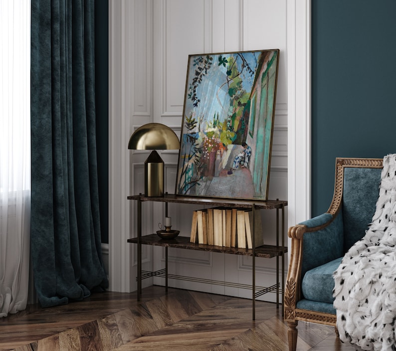 May include: A black metal shelf unit with a painting of a garden scene hanging above it. The shelf unit has three shelves and is standing on a wood floor. A gold table lamp is on the top shelf.