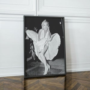 May include: Black and white photograph of a woman in a white dress with her skirt blowing in the wind. She is smiling and looking at the camera. The photo is framed in a black frame.