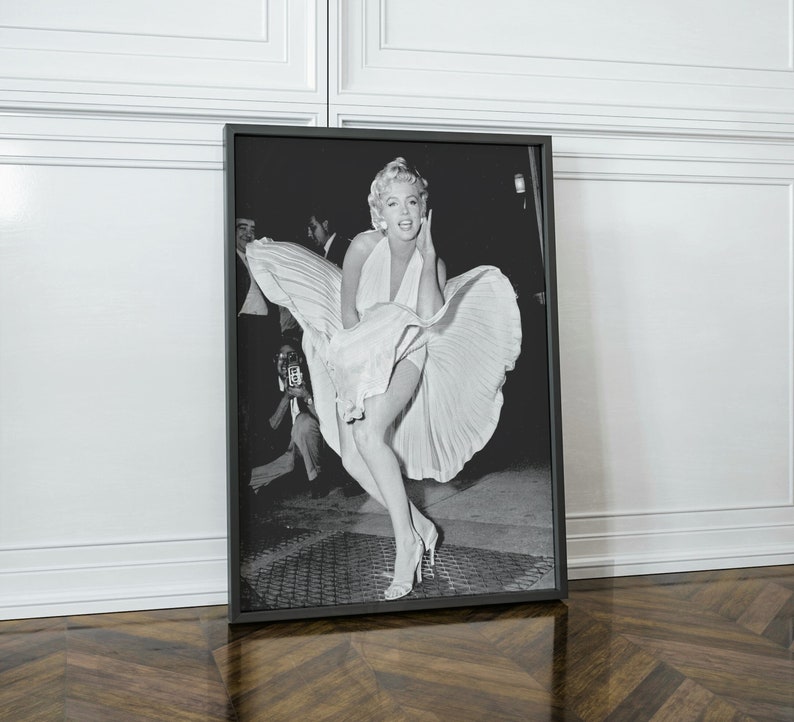 May include: Black and white photograph of a woman in a white dress with her skirt blowing in the wind. She is smiling and looking at the camera. The photo is framed in a black frame.