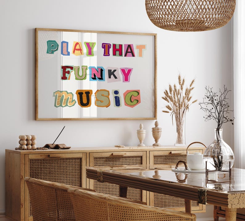 Puede incluir: Una impresi&oacute;n enmarcada con el texto "PLAY THAT FUNKY MUSIC" en letras de colores recortadas. La impresi&oacute;n est&aacute; colgada en una pared blanca sobre un armario de madera con puertas de rat&aacute;n tejido.