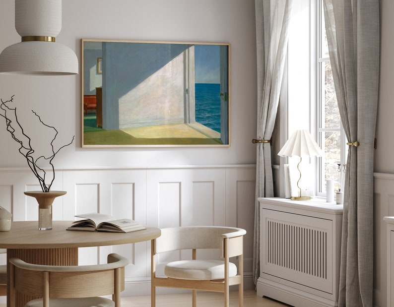 May include: A white dining room with a wooden table and chairs. A framed painting of a room with an open door leading to a view of the sea hangs on the wall. A white lamp with a white shade sits on a white radiator cabinet next to a window with white curtains.