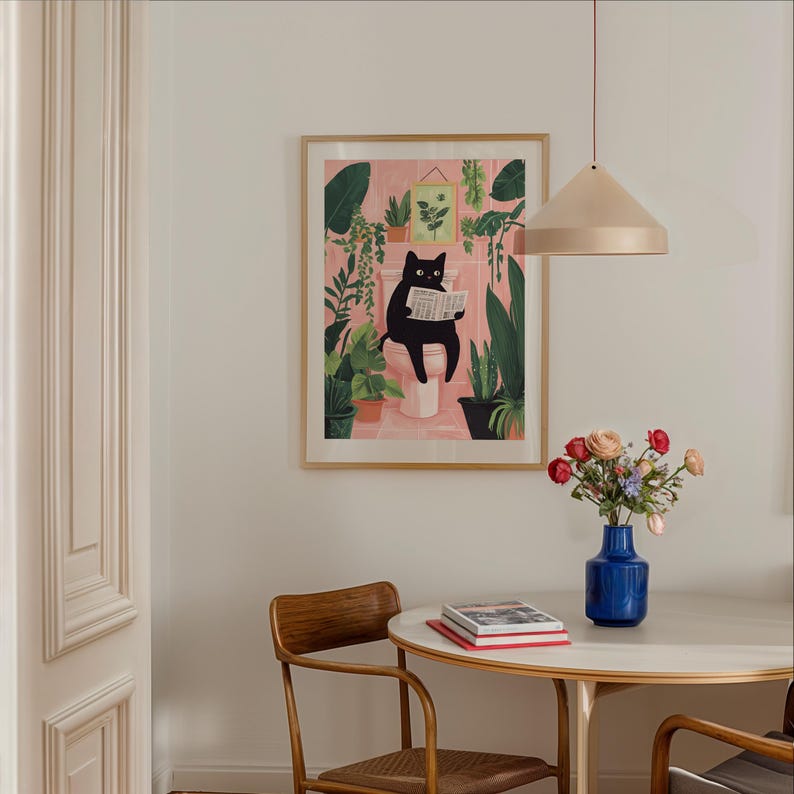 May include: A framed illustration of a black cat sitting on a toilet and reading a newspaper, surrounded by plants in a pink bathroom. The illustration is hanging on a white wall above a round wooden table with a vase of flowers and books.