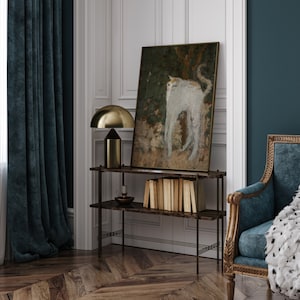 May include: A dark blue velvet curtain hangs in front of a window. A gold metal table lamp with a white shade sits on a black metal shelf. A painting of a white cat with a brown background hangs on the wall. A stack of books sits on the shelf. A dark blue velvet armchair with a white fur throw sits in the corner of the room.