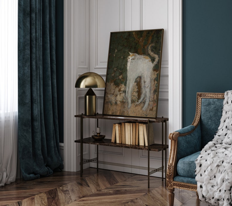 May include: A dark blue velvet curtain hangs in front of a window. A gold metal table lamp with a white shade sits on a black metal shelf. A painting of a white cat with a brown background hangs on the wall. A stack of books sits on the shelf. A dark blue velvet armchair with a white fur throw sits in the corner of the room.