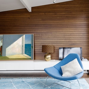 May include: A blue armchair with a white cushion sits in a living room with a wood-panelled wall, a white entertainment unit, and a painting of a seascape.