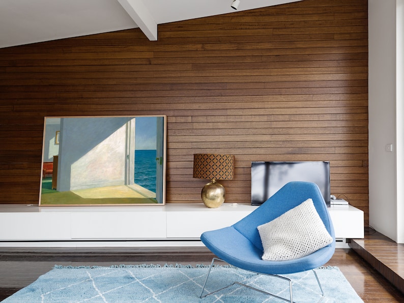 May include: A blue armchair with a white cushion sits in a living room with a wood-panelled wall, a white entertainment unit, and a painting of a seascape.