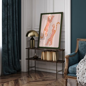 May include: A framed print of two pink and orange leopards leaping in the air against a light pink background. The frame is dark green and the print is hanging on a white wall.