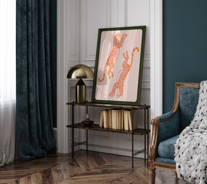 May include: A framed print of two pink and orange leopards leaping in the air against a light pink background. The frame is dark green and the print is hanging on a white wall.
