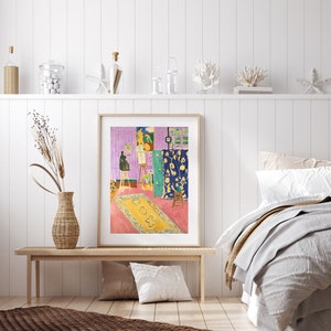 May include: A framed print of a painting depicting a room with a colorful rug, a green door, and a person painting on a canvas. The room is decorated with a blue and yellow patterned curtain.
