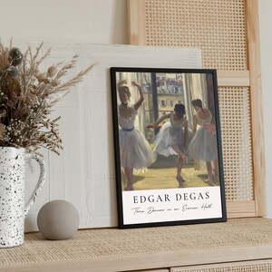 May include: A black framed print of a painting by Edgar Degas titled "Three Dancers in an Exercise Hall". The painting depicts three ballerinas in white tutus practicing in a dance studio.