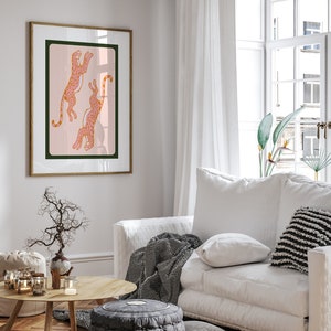 May include: A framed print of two pink leopards with orange spots jumping in the air against a light pink background. The frame is a light wood color.