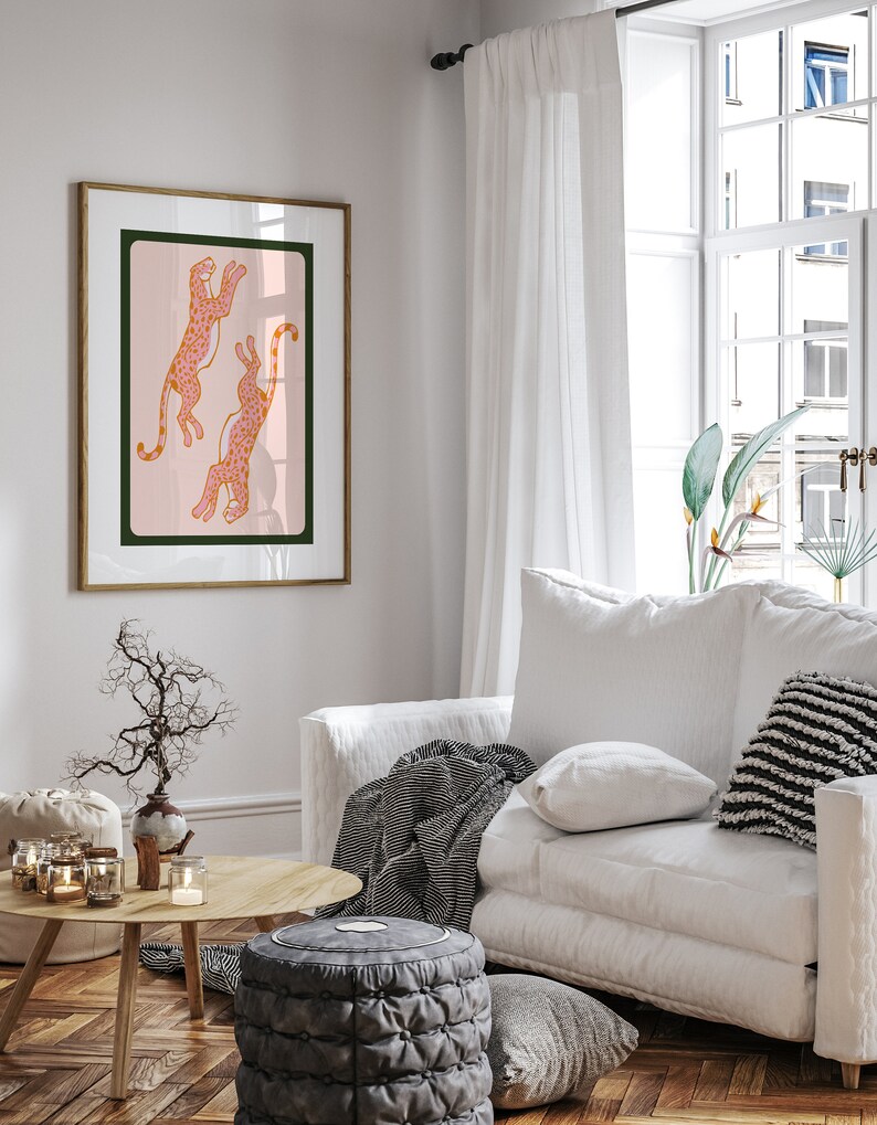 May include: A framed print of two pink leopards with orange spots jumping in the air against a light pink background. The frame is a light wood color.