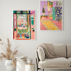 May include: Two framed paintings of abstract art in a living room setting. The painting on the left shows a view of a harbor with boats and a pink and green building. The painting on the right shows a room with a purple wall, a yellow rug, and a figure painting on an easel.