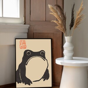 May include: Black and white illustration of a frog on a beige background with a red stamp in the top left corner. The illustration is framed in black and hanging on a wall.