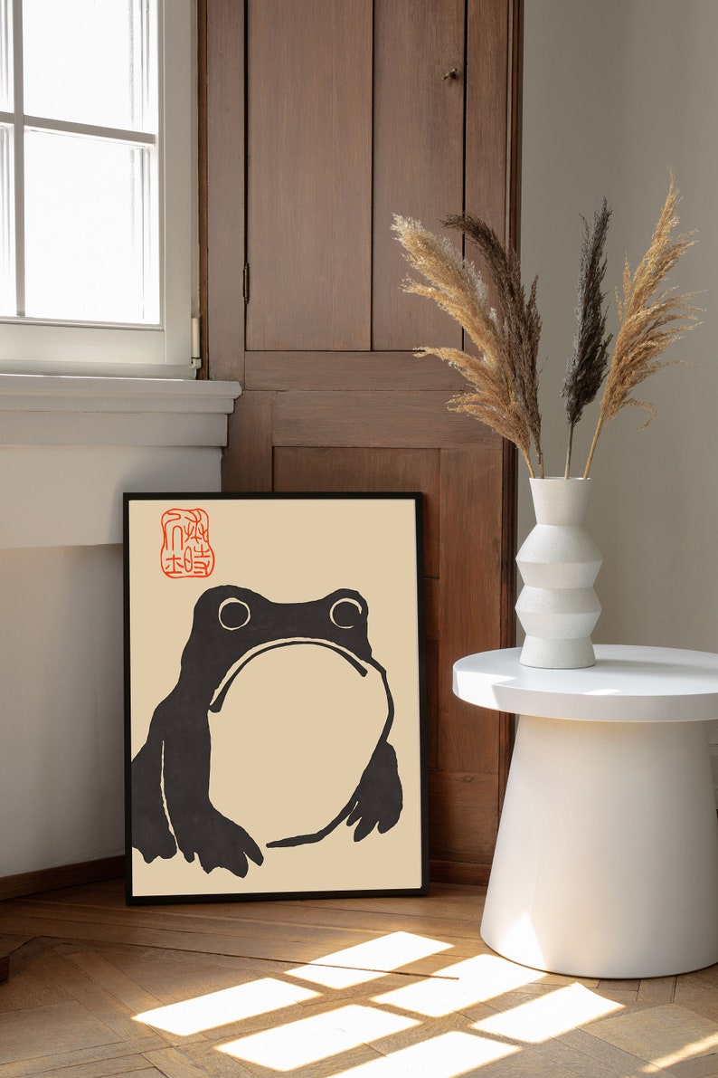 May include: Black and white illustration of a frog on a beige background with a red stamp in the top left corner. The illustration is framed in black and hanging on a wall.
