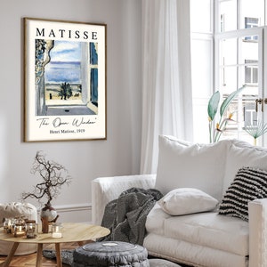 May include: A framed print of a painting by Henri Matisse titled "The Open Window". The painting depicts a view from a window with a blue sky, a palm tree, and a building in the distance. The print is in a white frame with a gold border.
