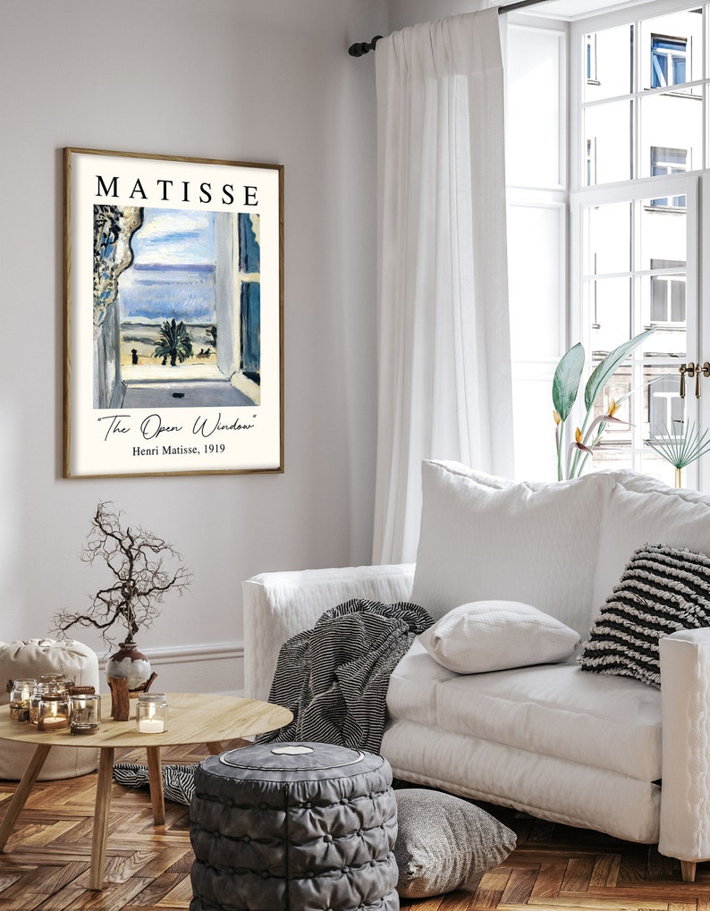 May include: A framed print of a painting by Henri Matisse titled "The Open Window". The painting depicts a view from a window with a blue sky, a palm tree, and a building in the distance. The print is in a white frame with a gold border.