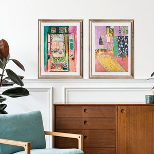 May include: Two framed art prints of paintings by Henri Matisse. The first print shows a view from a window with a pink wall and a green door. The second print shows a room with a purple wall, a yellow rug, and an easel with a painting on it.