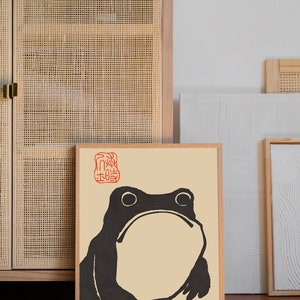 May include: A framed print of a black and white illustration of a frog with a red Japanese character stamp in the top left corner. The print is on a beige background and has a wooden frame.