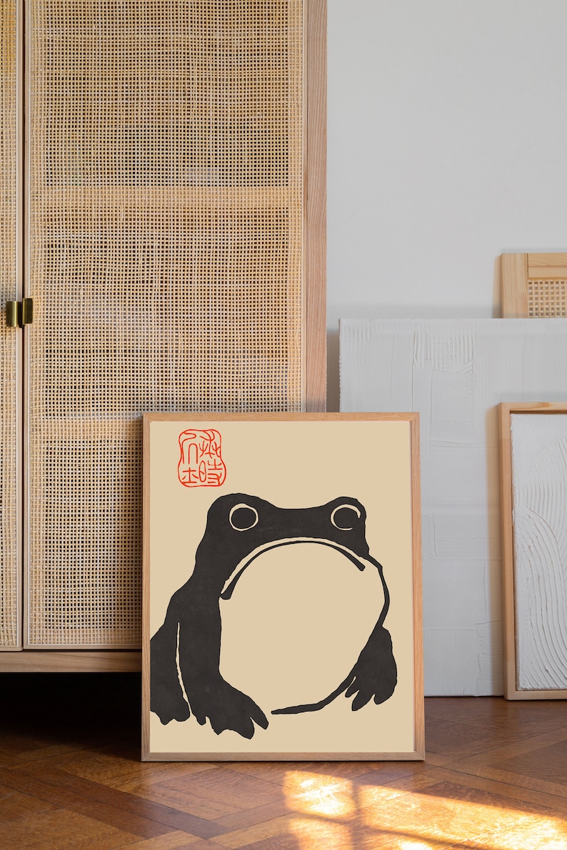 May include: A framed print of a black and white illustration of a frog with a red Japanese character stamp in the top left corner. The print is on a beige background and has a wooden frame.